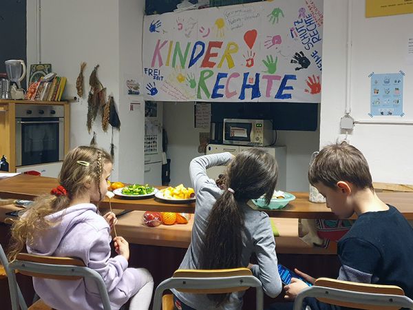 Drei Kinder essen eine Jause mit einem bunten "Kinderrechte" Plakatt im Hintergrund