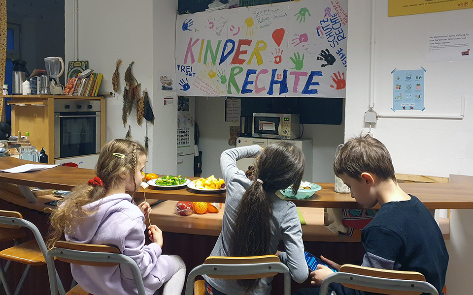 Jause Drei Kinder essen eine Jause mit einem bunten "Kinderrechte" Plakatt im Hintergrund